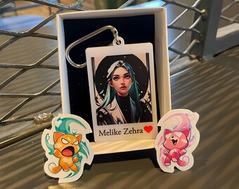 Op de afbeelding: Een sleutelhanger met een portret van een vrouw met turquoise haar en de naam "Melike Zehra". Er zijn ook twee kattenstickers, één oranje en één roze. De items worden gepresenteerd in een witte doos.