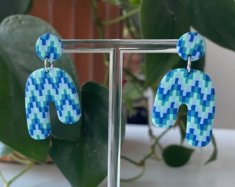 May include: A pair of blue and green geometric patterned earrings in the shape of an arch. The earrings are displayed on a clear stand.