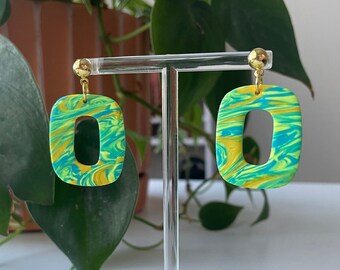 May include: A pair of green and yellow swirl patterned earrings with gold hooks. The earrings are displayed on a clear stand.