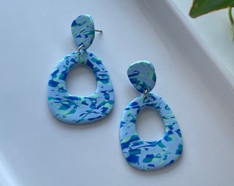 May include: A pair of handmade polymer clay earrings. The earrings feature a light blue base with a speckled pattern of dark blue, teal, and green. Each earring has a small, rounded stud and a larger, open teardrop shape.