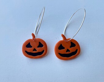 May include: A pair of Halloween-themed earrings. Each earring features a small, orange pumpkin with a carved face in black. The pumpkins are suspended from thin, silver-colored hoop wires.