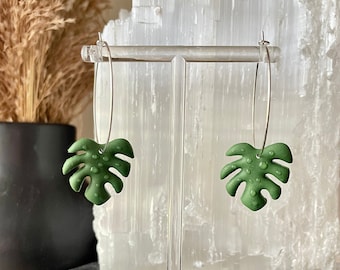 May include: A pair of silver hoop earrings with green monstera leaf charms. The leaves have a textured, dotted surface.