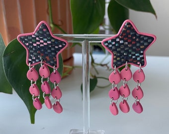 May include: A pair of pink and black star-shaped earrings with dangling pink beads. The earrings are displayed on a clear stand.
