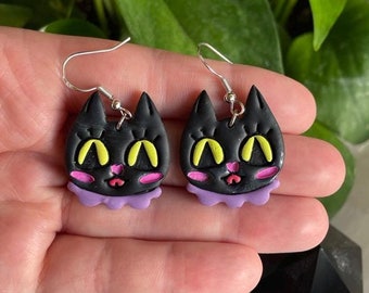 May include: A pair of black cat-shaped dangle earrings with yellow eyes, pink cheeks, and a purple ruffle. The earrings are made of polymer clay and have silver hooks.