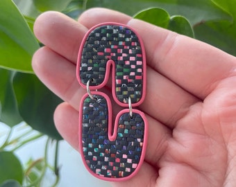 May include: A pair of pink and black statement earrings with a woven pattern. The earrings are shaped like the letter 'S' and have a pink border.