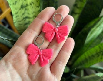 May include: A pair of bright pink bow-shaped earrings. The earrings are made of a textured material and have silver-colored leverback closures. The bows are a vibrant, solid pink color, and the earrings are displayed against a background of green foliage.