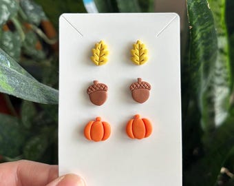 May include: Three pairs of stud earrings attached to a white display card. The top pair features yellow oak leaves, the middle pair consists of brown acorns, and the bottom pair displays orange pumpkins. Each earring has a smooth, matte finish.