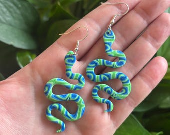May include: A pair of green and blue snake earrings with a swirling pattern. The earrings are made of polymer clay and have silver hooks.