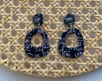 May include: A pair of black dangle earrings with a floral pattern and a silver hook. The earrings are oval shaped with a hole in the center.