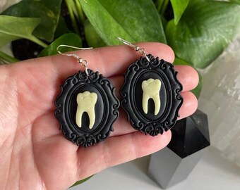 May include: A pair of dangle earrings featuring an oval black frame with ornate, raised scrollwork detailing. Centered within each black frame is a pale, off-white molar tooth shape. The earrings hang from silver-toned metal hooks.