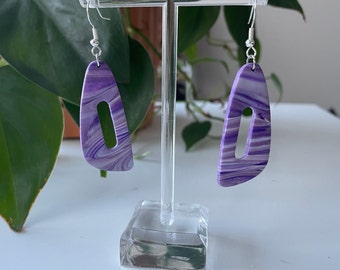 May include: A pair of purple and white marbled dangle earrings with a silver hook. The earrings are shaped like a teardrop with a cut-out oval in the center.