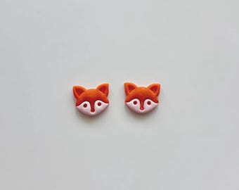 May include: A pair of stud earrings shaped like fox heads. Each earring features a bright orange upper portion with pointed ears and a light pink lower portion forming the muzzle and cheeks, with small circular indentations for the eyes and nose.