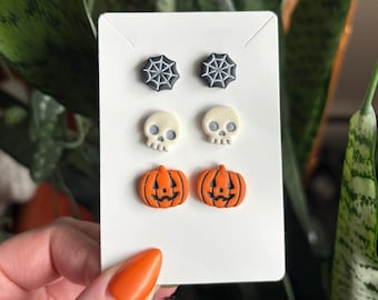 May include: Three pairs of stud earrings displayed on a white card. The top pair features black spiderwebs, the middle pair consists of off-white skulls, and the bottom pair depicts orange jack-o'-lantern pumpkins with black carved facial features.