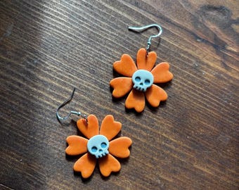 May include: A pair of dangle earrings featuring orange flower-shaped charms with six heart-shaped petals. Each flower has a light blue skull centered in the middle. The earrings are attached to silver-toned metal hooks for pierced ears.
