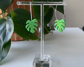 May include: A pair of green and yellow leaf-shaped earrings on a clear acrylic earring stand. The earrings are made of polymer clay and have a marbled effect.