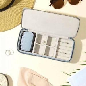 Puede incluir: Un estuche de joyería azul claro con cremallera, con compartimentos para anillos, pendientes y una pequeña bolsa. El estuche está abierto y el interior está forrado con un tejido blanco suave.