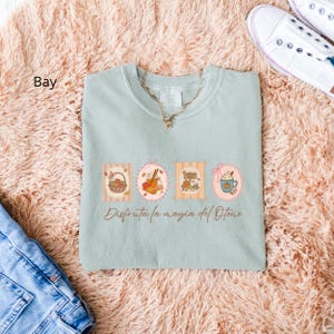 May include: A light green t-shirt with a gold necklace and a graphic of four fall-themed illustrations. The illustrations include a basket of fruit, a jar of apple cider, pumpkins, and a mug of coffee. The text "Disfruta la magia del Otoño" is below the illustrations.