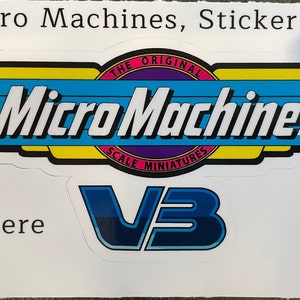 May include: A white sticker with a blue, yellow, and pink Micro Machines logo. The logo says "The Original Scale Miniatures" and has a blue and white wing design. The sticker also has a blue and white "V3" logo and says "Peel Here" with an arrow pointing up.