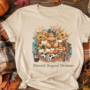 May include: A beige t-shirt featuring a graphic of a highland cow sitting in the back of a teal pickup truck filled with pumpkins, sunflowers, and wheat. The cow wears a patterned headband and necklace. The text Blessed Beyond Measure is below.