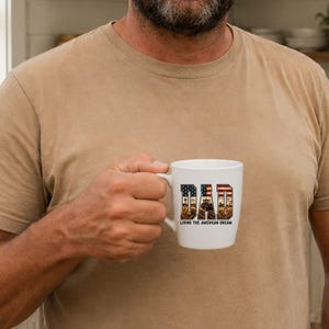 Puede incluir: Taza de cer&aacute;mica blanca con la palabra "DAD" en un dise&ntilde;o patri&oacute;tico con la bandera estadounidense y una escena rural. El texto "Living the American Dream" est&aacute; debajo de la palabra "DAD".