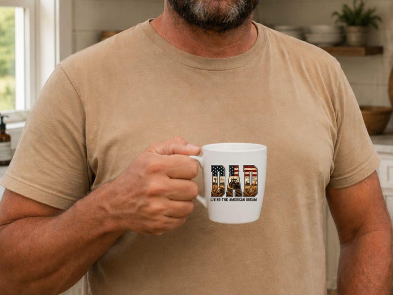 Puede incluir: Taza de cer&aacute;mica blanca con la palabra "DAD" en un dise&ntilde;o patri&oacute;tico con la bandera estadounidense y una escena rural. El texto "Living the American Dream" est&aacute; debajo de la palabra "DAD".