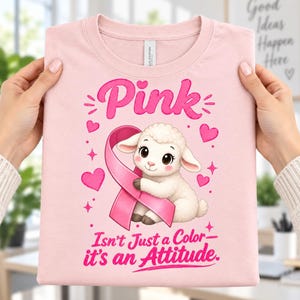 Puede incluir: Una camiseta rosa claro con un gráfico de un cordero blanco abrazando un lazo de concienciación rosa. El texto superior dice Pink y el texto inferior dice Isn't Just a Color—it's an Attitude. El diseño incluye varios corazones y estrellas.