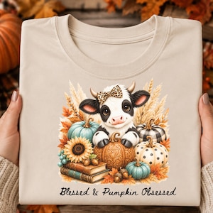 May include: A beige t-shirt featuring a graphic of a cow with a leopard print bow, surrounded by pumpkins, sunflowers, and books. The text "Blessed & Pumpkin Obsessed" is printed in black cursive script below the central illustration.