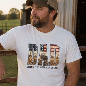 Puede incluir: Camiseta blanca con la palabra "DAD" en un dise&ntilde;o patri&oacute;tico, con la bandera estadounidense y un paisaje rural. El texto "LIVING THE AMERICAN DREAM" est&aacute; debajo de la palabra "DAD".