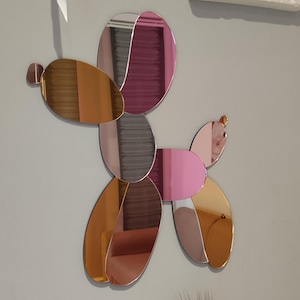 Könnte beinhalten: Wandskulptur in Form eines Ballonhundes. Die Skulptur besteht aus verspiegeltem Acryl in Gold-, Rosa-, Grau- und Burgundertönen. Der Hund besteht aus mehreren ovalen und runden Formen.