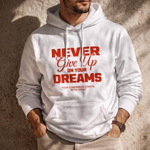 Könnte beinhalten: Weißer Kapuzenpullover mit Fronttasche und Kordelzug. Der rote Text "NEVER Give Up ON YOUR DREAMS" und "YOUR TOMORROW STARTS WITH TODAY" ist auf der Vorderseite aufgedruckt. Eine braune Uhr ist sichtbar.