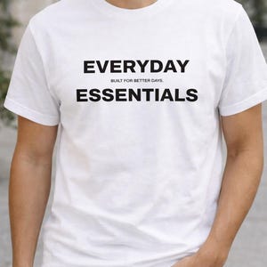 Könnte beinhalten: Weißes T-Shirt mit den Wörtern "EVERYDAY ESSENTIALS" in Schwarz aufgedruckt, mit dem Satz "BUILT FOR BETTER DAYS" über dem Wort "ESSENTIALS". Das Shirt hat kurze Ärmel.