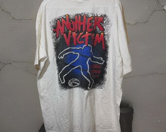May include: White t-shirt with a graphic on the back. The graphic features the words "ANOTHER VICTIM" in red, a blue outline of a body, and other text. The design is set against a black background.