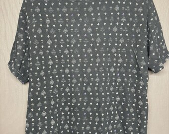 May include: Dark gray t-shirt with an all-over pattern of white spades, skulls, and purple accents. The word "Vurt" is printed in purple on the neckline. The short-sleeved shirt is hanging on a black hanger.