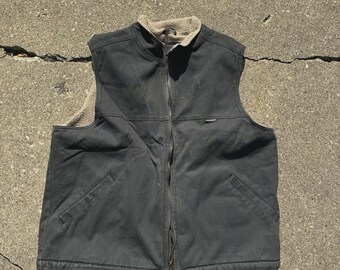 May include: A dark gray vest with a full-length zipper and a tan, fuzzy collar. The vest has two front pockets and a small logo on the chest. The vest appears to be made of a durable, weather-resistant material.
