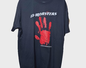 May include: Black t-shirt with the text "13-MONSTERS" in white and a large red handprint graphic. The text "13MONSTERSMUSIC.COM" is printed below the handprint. The shirt is hanging on a white hanger.