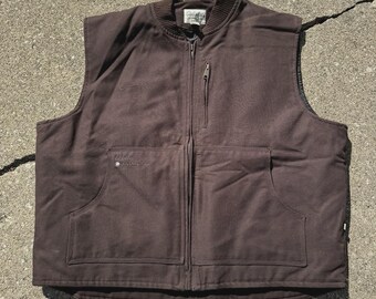 May include: A brown vest with a zip-up front, two large front pockets, and a ribbed collar. The vest has a small logo on the chest and a tag on the collar. The vest is sleeveless and appears to be made of a durable material.