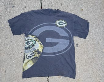 May include: Dark gray t-shirt featuring the Green Bay Packers logo and a championship ring graphic. The shirt has a large green and gray "G" logo on the front, with a smaller logo on the upper right. The championship ring graphic is on the left side.