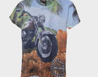 May include: A light blue t-shirt featuring a motorcycle graphic with a tropical background. The Harley-Davidson logo is visible on the lower right. The shirt is hanging on a white hanger.