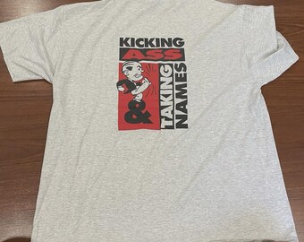 May include: A light gray t-shirt with the words "KICKING ASS & TAKING NAMES" in black and red. A cartoon figure is in the center of the design. The shirt is laid flat on a wooden surface.