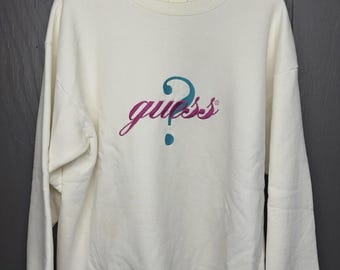 May include: Off-white sweatshirt with the word "guess" in a script font, with a question mark above the word. The text is in a gradient of purple and teal. The sweatshirt is hanging on a metal hanger.