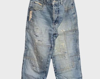May include: Light blue denim jeans with a patchwork design. The jeans have a faded wash and distressed details, including rips and patches. The jeans are hanging on a clear hanger.