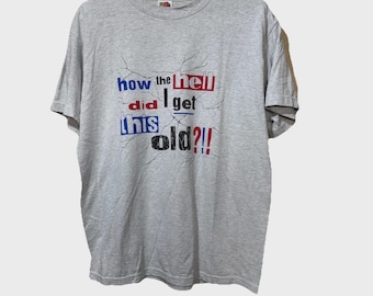 May include: A light gray t-shirt with the text "how the hell did I get this old?!" in red, blue, and white. The text has a cracked effect. The shirt is hanging on a white hanger.