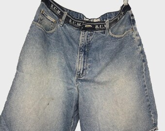 May include: Light blue denim shorts with a black waistband featuring the text "B.U.M. Equipment" in white. The shorts have a classic five-pocket design and a faded wash. The shorts are hanging from a metal clip.