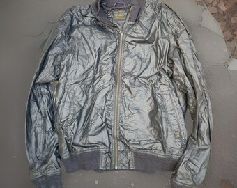 May include: A metallic silver bomber jacket with a full zipper closure. The jacket has a ribbed collar, cuffs, and hem in a dark gray color. The jacket has a shiny, crinkled texture and a small tag on the chest.