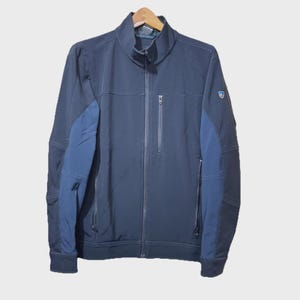 May include: A navy blue jacket with a full-length zipper and a stand-up collar. The jacket features dark blue panels on the sleeves and a small logo on the upper arm. It has two zippered pockets and a chest pocket.