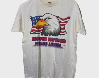May include: Cream-colored t-shirt featuring a graphic of an American bald eagle with a red, white, and blue American flag in the background. The text "WE SUPPORT OUR TROOPS GOD BLESS AMERICA" is printed below the eagle.