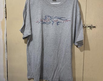 May include: A gray t-shirt with a crew neck and short sleeves. The shirt features a tribal design in red, white, and blue across the chest. The shirt is hanging on a white hanger.