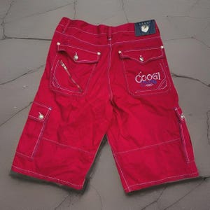 May include: Red cargo shorts with white stitching and multiple pockets. Features a zipper pocket and the "Coogi" logo embroidered in blue. Includes a small blue tag with the brand name. The shorts are laid flat.