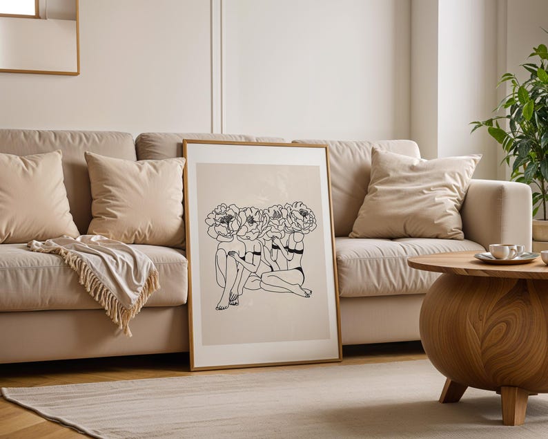 Puede incluir: Una impresi&oacute;n art&iacute;stica enmarcada con un dibujo lineal minimalista de tres figuras con cabezas florales. La obra de arte est&aacute; en un marco dorado y se apoya en un sof&aacute; beige con cojines a juego. Una mesa de centro de madera y una alfombra est&aacute;n en primer plano.