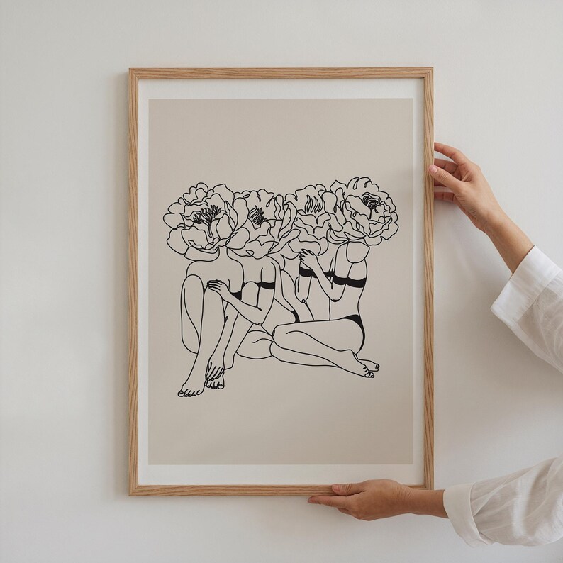 Puede incluir: Una impresi&oacute;n art&iacute;stica enmarcada con un dibujo lineal minimalista de tres figuras con cabezas de flores de peon&iacute;a. La obra de arte en blanco y negro est&aacute; sobre un fondo beige, sostenida por manos con una camisa blanca.